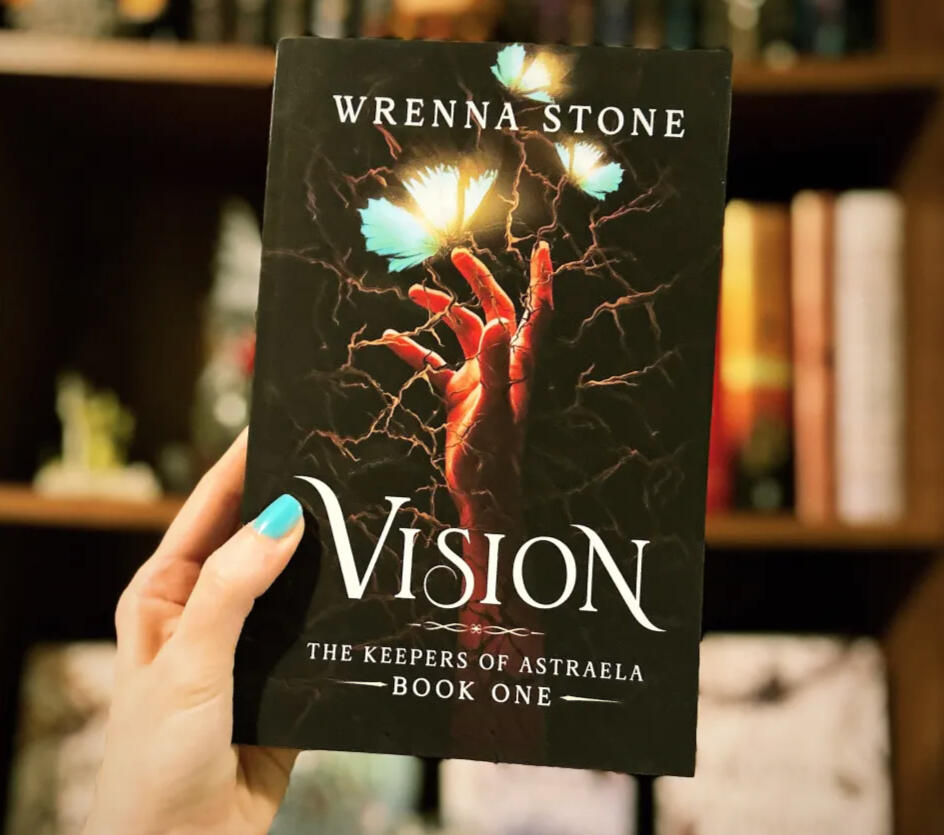 A hand holding up the novel "Vision," the first book in the Keepers of Astraela series. The book cover features a dark scheme with vines behind a hand reaching towards luminescent blue moths.